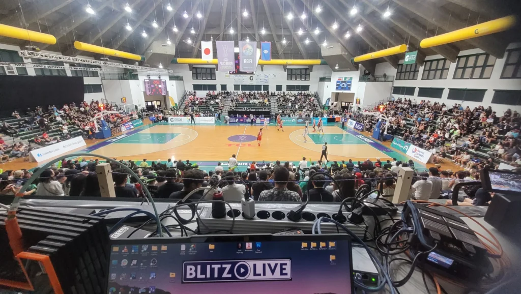 A wide-angle view of a basketball arena filled with fans. The image is taken from a production booth, showing multiple equipment setups, including a monitor displaying the "BLITZ LIVE" logo